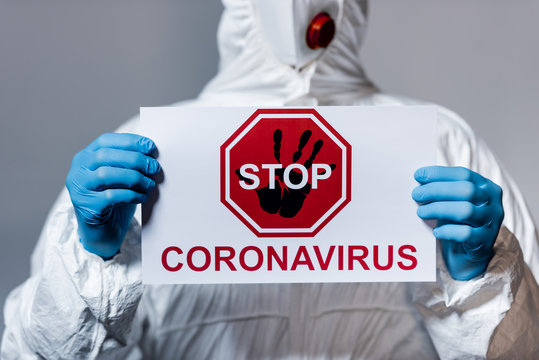 Cropped View Of Mature Doctor In Hazmat Suit Holding Placard With Stop Coronavirus Lettering Isolated On Grey