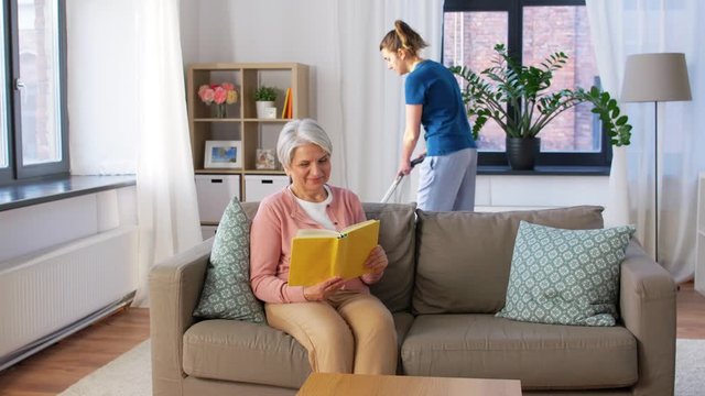 leisure, old age and people concept - happy senior woman reading book and housekeeper cleaning at home