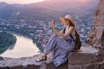 Beautiful young woman in long dress and hat, sits on hill and photographing the landscape on phone.