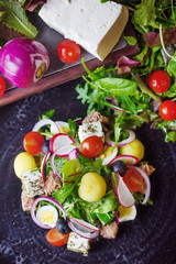 A salad of cherry tomatoes,arugula,lots of herbs,radishes,eggs,cheese and some meat.All the ingridients are on a dark table.
