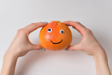 Grapefruit with funny face in female hands on white background. Orange with Googly eyes and painted smile