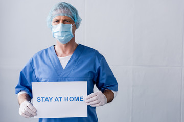 mature doctor in medical mask, cap and latex gloves holding placard with stay at home lettering on grey