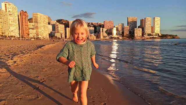 Blonde 3-years-old girl in knitted sweater starts running along the city's sandy beach at sunset. Slow motion.