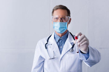 selective focus of mature doctor in medical mask and googles holding test tube with blood sample on...