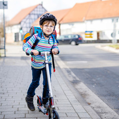 Active school kid boy in safety helmet riding with his scooter in the city with backpack on sunny day. Happy child in colorful clothes biking on way to school. Safe way for kids outdoors to school