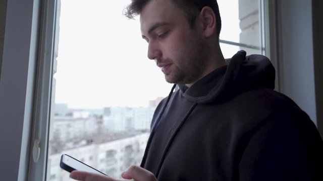 Amazed young man checks his phone, smoking iqos by the window, close up