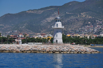 Fototapeta na wymiar Beautiful lighthouse on the sea shore. A view of the city on the mountain under clear blue skies.