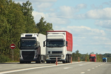 Two oncoming European semi trucks overtaking, vehicle front side view on suburban highway road on summer day on trees and blue sky background