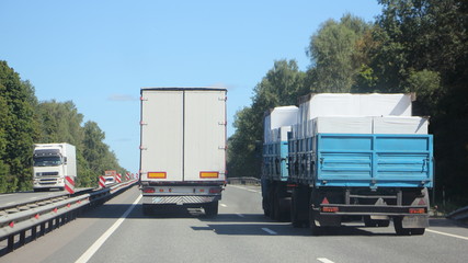 Semi truck with white van trailer overtakes loaded flatbed truck on suburban highway, rear view from road at summer day on clear blue sky and forest background, cargo logistics