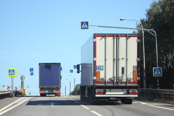White and blue semi trucks rear view on Sunny summer suburban highway on blue sky background, goods delivery with road cargo transportation logistics in Europe