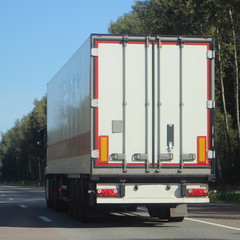 White European semi truck drive on suburban Sunny summer highway road, rear side view in perspective on blue sky background, contemporary logistic business, international cargo transportation industry