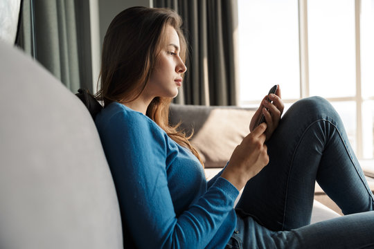 Image of young serious woman using mobile phone while sitting on sofa