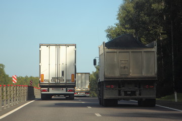 White semi truck with van trailer overtake loaded flatbed dump truck on summer country highway road, rear view on trees and blue sky background
