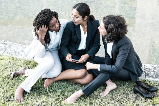 High angle view of businesswomen using smartphone outdoors. Group of professional multiethnic businesswomen sitting on lawn and using smartphone together. Technology concept