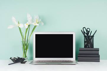 Elegant spring home workplace with blank notebook screen, black stationery, books, white fresh flowers in soft light green mint menthe interior on white wood desk.