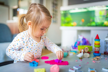 Happy little child, adorable creative 2 year old girl playing with dough, colorful modeling compound, sitting bright sunny room at home.
