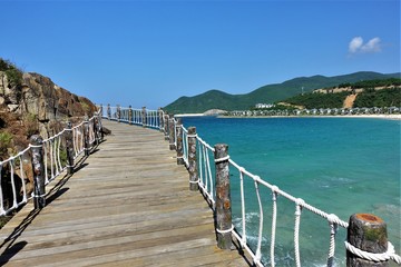 Summer. Sunny day. A wooden path with rope handrails goes over the turquoise sea around the mountain into the unknown. In the distance are green hills, a white beach and small houses.