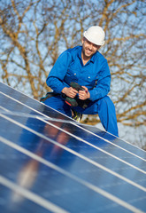 Portrait of man worker in blue suit and protective helmet installing stand-alone solar photovoltaic panel system using screwdriver. Smiling electrician mounting blue solar module on roof of house.