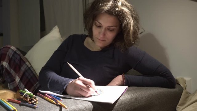 Footage of caucasian woman painting on paper with coloured pencils on a couch at home. Medium slide shot