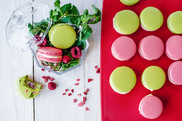 Colorful French macaroons dessert in the glass vase near red boar with macarons cookie on white wooden background.The composition of food is decorated with raspberries, mint leaves.Flat lay with copy 