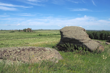 The landscape with the big grey stones, the field with green grass and flowers, the far rock, the white clouds in the blue sky.
