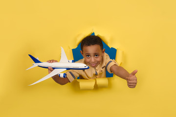 African-American boy smiles and looks out of wall of torn paper on yellow, holds airplane in his hands and shows finger up, approves airlines or travel. Concept tourism air travel and summer holidays