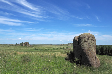The landscape with the big grey stone, the field with green grass and flowers, the far rock, the white clouds in the blue sky.