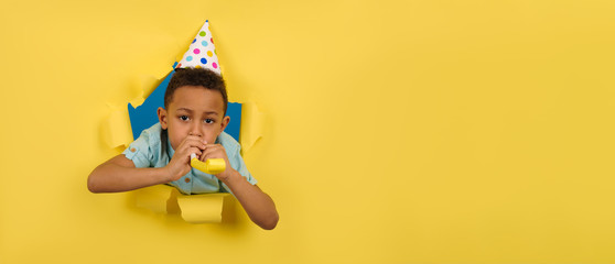 happy Afro American boy blowing festive pipe at birthday party with a cone cap on his head on a...