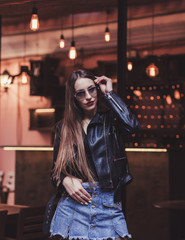 Trendy girl in sunglasses and leather jacket posing in evening city