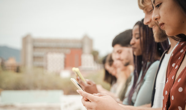 Teenagers texting mobile phone messages sitting on urban wall - Group of friends millennial using cellular outdoors - Concept of social network and telephone technology. Image