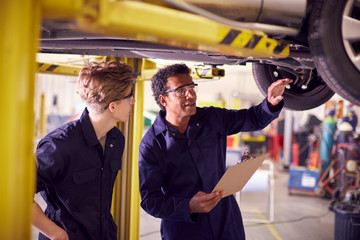 Male Tutor With Student Looking Underneath Car On Hydraulic Ramp On Auto Mechanic Course 