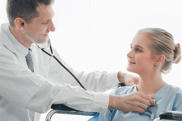 Doctor holding stethoscope for doing a physical examination of the young beautiful patient who setting on a wheelchair in medical office, health care and hospital concept