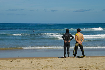Two surfer guys stand on sand in wetsuits and look at the waves. Bay of Biscay. Surfing in Spain.