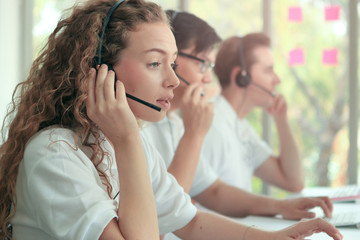 Young beautiful woman with headphones working at call center service desk consultant with her teammates, talking with the customer on hands-free phone