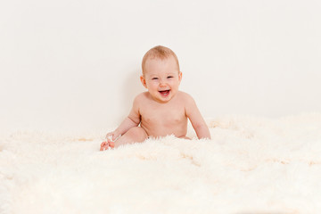 a naked Caucasian eight-month-old baby sits on a bed against a white background and laughs.