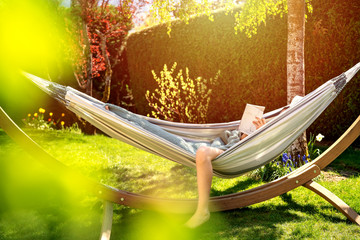 Young relaxed girl reading book in hammock in garden at home at bright sunset. Slow living, gadget...