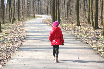 Little girl in red jacket and red hat walks in spring forest among trees on asphalt road