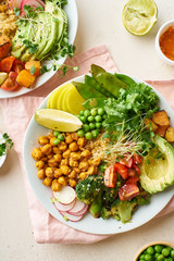 Healthy vegetarian lunch bowl with avocado, chickpeas, quinoa and vegetables, garnished with microgreens and nut dressing. Flat lay on stone background.