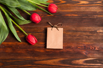 Tiny paper gift pack near fresh red tulips with green leaves on dark wooden background