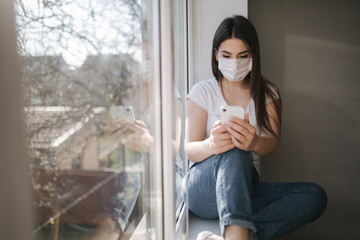 Attractive woman in medical mask sits on windowsill at home. Female in white t'shirt and white medical mask use phone