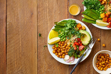 Healthy vegetarian lunch bowl with avocado, chickpeas, quinoa and vegetables, garnished with microgreens and nut dressing. Flat lay on wooden background.