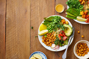 Healthy vegetarian lunch bowl with avocado, chickpeas, quinoa and vegetables, garnished with microgreens and nut dressing. Flat lay on wooden background.