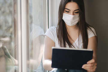 Attractive young woman in white medical mask sits on windowsiil and use tablet. Female have a rest at home in quarantine