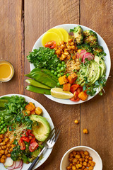 Healthy vegetarian lunch bowl with avocado, chickpeas, quinoa and vegetables, garnished with microgreens and nut dressing. Flat lay on wooden background.