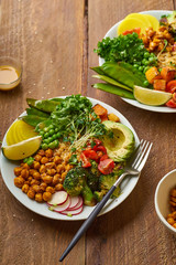 Healthy vegetarian lunch bowl with avocado, chickpeas, quinoa and vegetables, garnished with microgreens and nut dressing. Flat lay on wooden background.