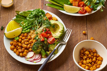 Overhead shot of healthy vegetarian lunch bowl with avocado, chickpeas, quinoa and vegetables, garnished with microgreens and nut dressing. Flat lay on wooden background.