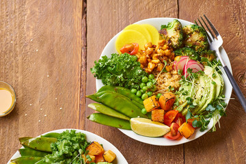 Healthy vegetarian lunch bowl with avocado, chickpeas, quinoa and vegetables, garnished with microgreens and nut dressing. Flat lay on wooden background.