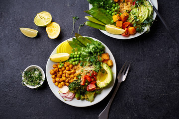 Flat lay with healthy vegetarian lunch bowl with avocado, chickpeas, quinoa and vegetables, garnished with microgreens and nut dressing. Dark concrete background, copy space.