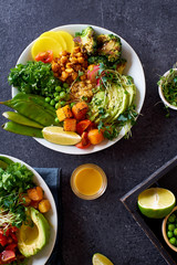 Healthy vegetarian lunch bowl with avocado, chickpeas, quinoa and vegetables, garnished with microgreens and nut dressing. Flat lay on dark concrete background.
