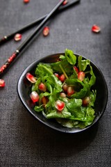 Closeup of arugula salad with pomegranate seeds in black dish on dark gray background with some chopsticks in the background. Macro, vertical, shallow DOF.
